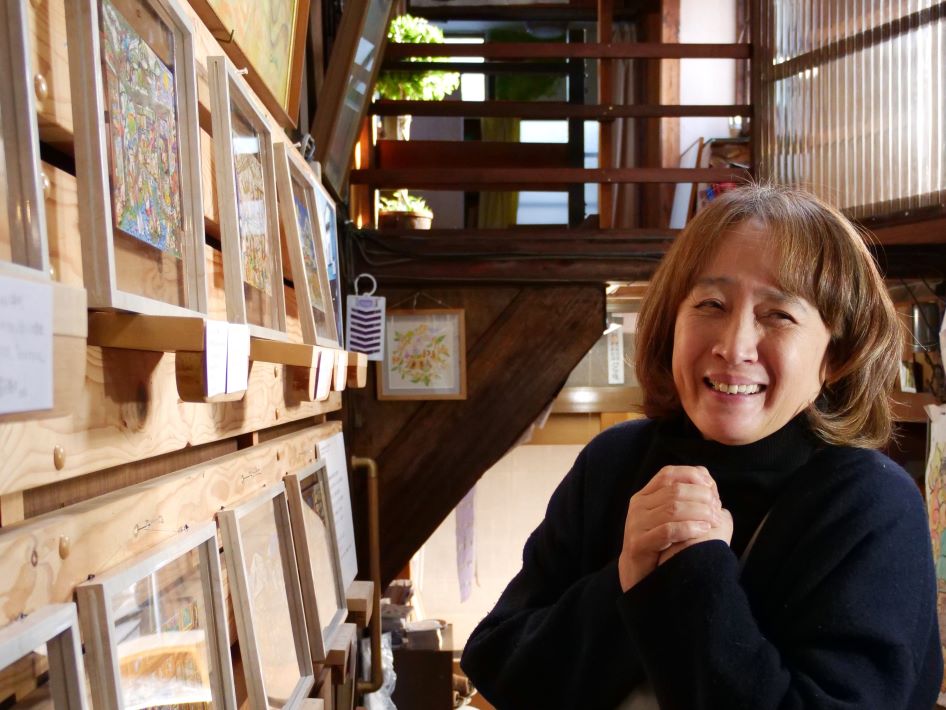 「100年生きられた家」の原画の前で話をする店主の佐藤真里さん