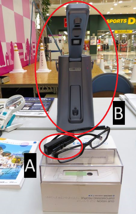 視覚障害「ロービジョン」を知る商品体験会 A眼鏡フレームに装着できる小型カ メラとスピーカー搭載の活字文書読み上げ装置。B印刷された文字を撮影して音声で読み上げる音声読書器