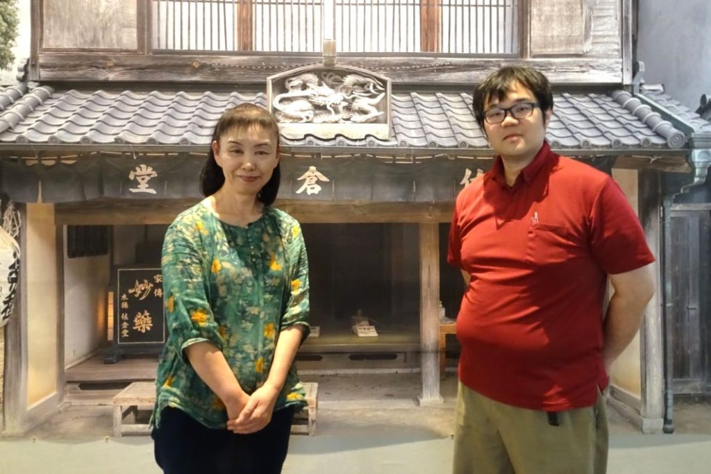 「生薬～自然からの恵み～」薬屋「佐倉堂」の写真パネルの前に立つ企画 展担当者